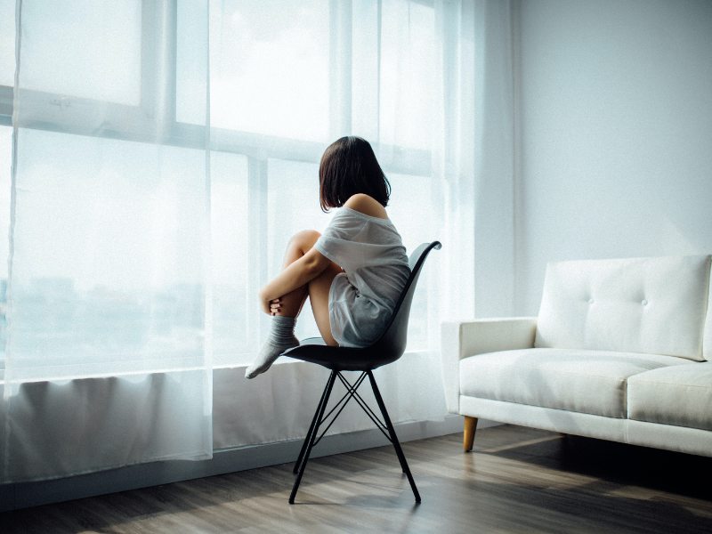 Female student sitting on chair facing window in living room holding legs and looking out of window 